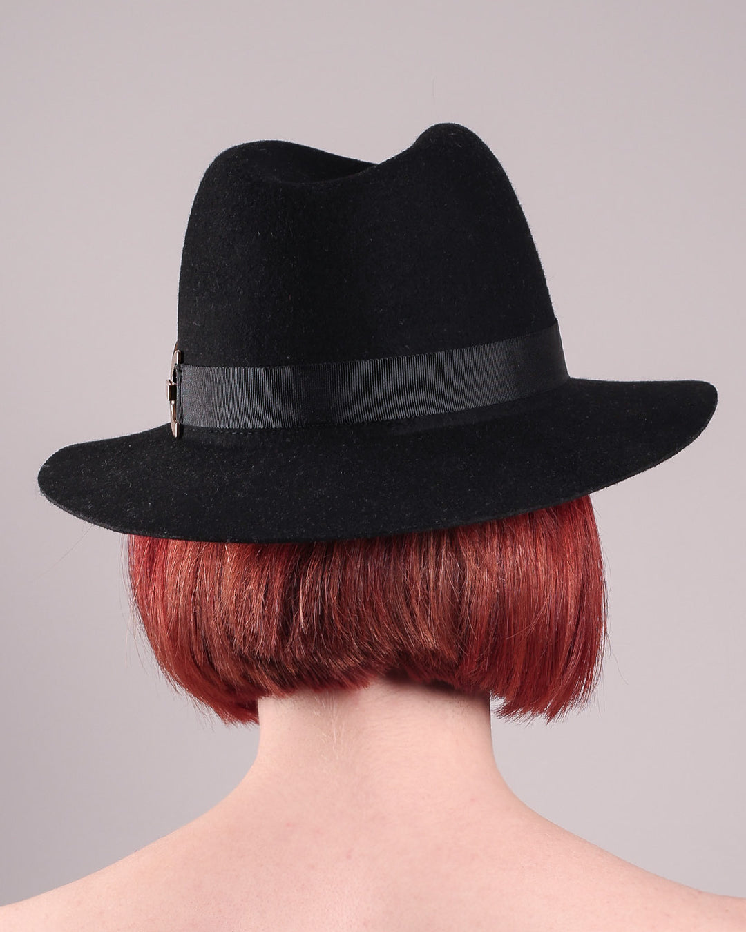 The model showcases the back of the Nocturne Fedora hat, crafted from rich black wool felt. This structured silhouette is elevated by a gold coloured, bold metallic chain detail in warm gold on the left side, linked seamlessly into a sleek black grosgrain band.