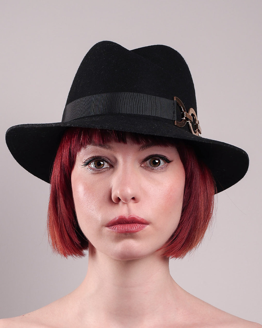 The model showcases the front close up of the Nocturne Fedora hat, crafted from rich black wool felt. This structured silhouette is elevated by a gold coloured, bold metallic chain detail in warm gold on the left side, linked seamlessly into a sleek black grosgrain band.