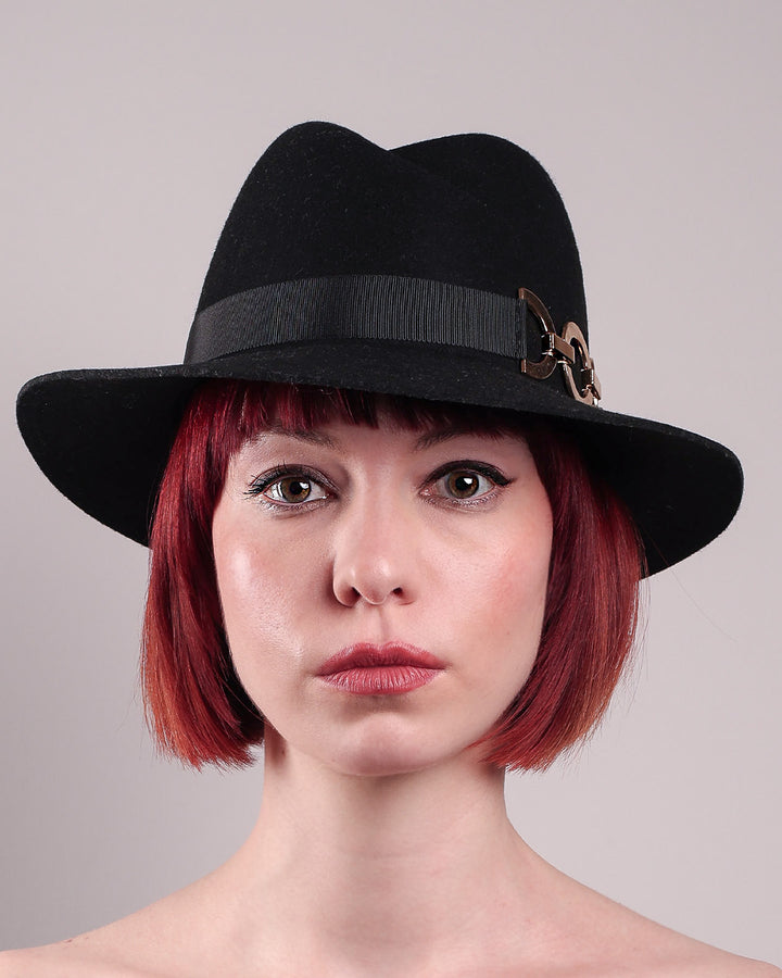 The model showcases the front close up of the Nocturne Fedora hat, crafted from rich black wool felt. This structured silhouette is elevated by a gold coloured, bold metallic chain detail in warm gold on the left side, linked seamlessly into a sleek black grosgrain band.