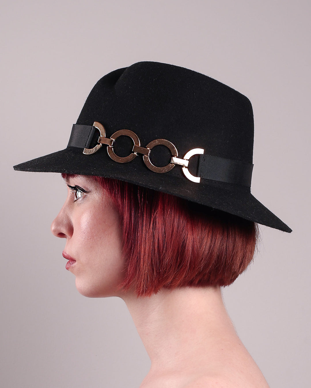 The model showcases the left close up of the Nocturne Fedora hat, crafted from rich black wool felt. This structured silhouette is elevated by a gold coloured, bold metallic chain detail in warm gold on the left side, linked seamlessly into a sleek black grosgrain band.