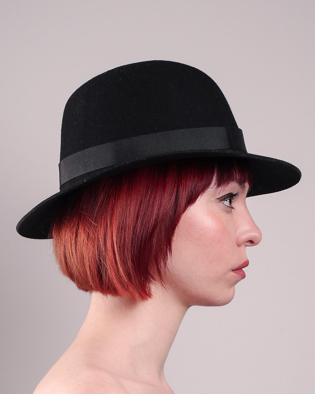 The model showcases the right side close up of the Nocturne Fedora hat, crafted from rich black wool felt. This structured silhouette is elevated by a gold coloured, bold metallic chain detail in warm gold on the left side, linked seamlessly into a sleek black grosgrain band.