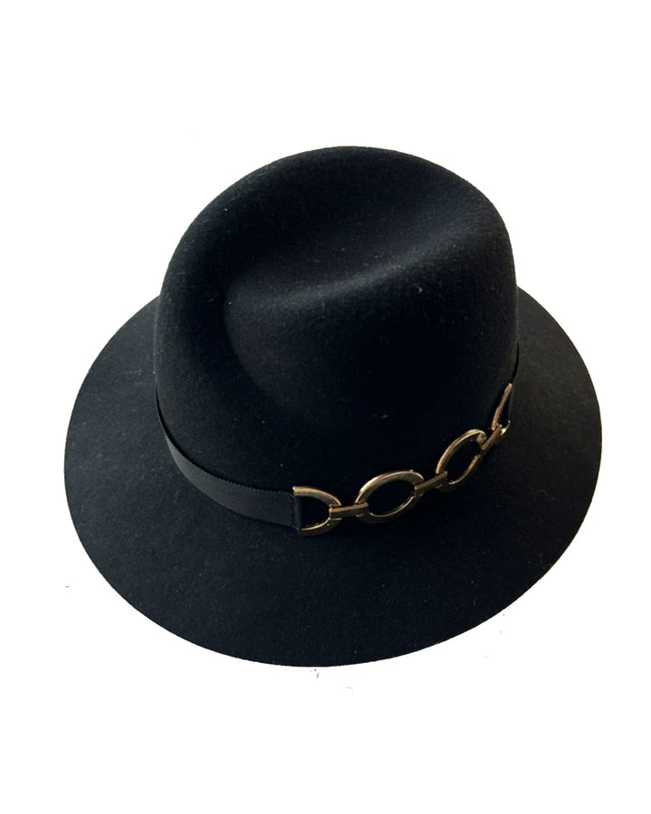 Still life picture of the Nocturne Fedora hat, crafted from rich black wool felt. This structured silhouette is elevated by a gold coloured, bold metallic chain detail in warm gold on the left side, linked seamlessly into a sleek black grosgrain band.