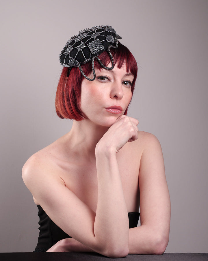 The model showcases the right side of the Nova Pillbox Fascinator is an intricate constellation of geometry and texture. Its classic pillbox base in black wool felt is overlaid with a meticulously hand-stitched web of silver thread, creating a dimension by adding the formed rhinestone pipe grid that breaks into flowing strands at the edges. Designed by Merve Bayindir.