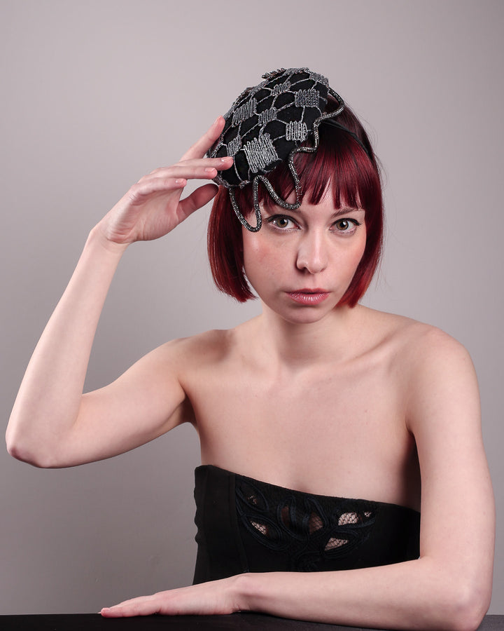 The model showcases the front higher angle of the Nova Pillbox Fascinator is an intricate constellation of geometry and texture. Its classic pillbox base in black wool felt is overlaid with a meticulously hand-stitched web of silver thread, creating a dimension by adding the formed rhinestone pipe grid that breaks into flowing strands at the edges. Designed by Merve Bayindir.