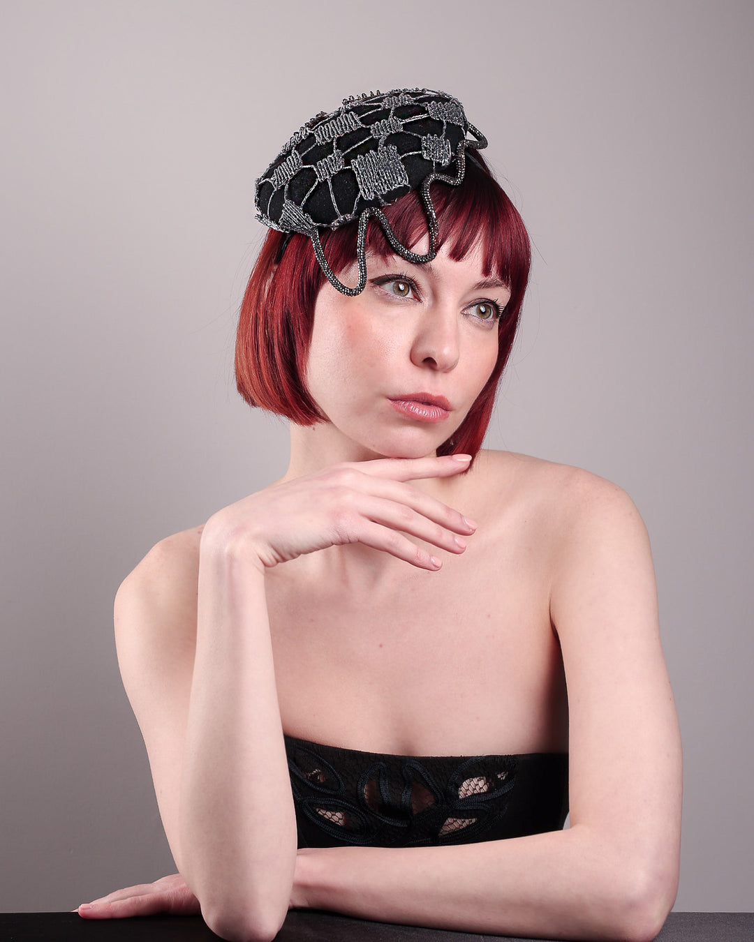 The model showcases the front right angle of the Nova Pillbox Fascinator is an intricate constellation of geometry and texture. Its classic pillbox base in black wool felt is overlaid with a meticulously hand-stitched web of silver thread, creating a dimension by adding the formed rhinestone pipe grid that breaks into flowing strands at the edges. Designed by Merve Bayindir.