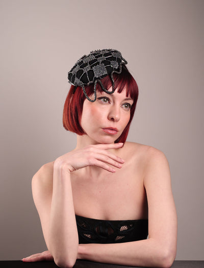 The model showcases the front right angle of the Nova Pillbox Fascinator is an intricate constellation of geometry and texture. Its classic pillbox base in black wool felt is overlaid with a meticulously hand-stitched web of silver thread, creating a dimension by adding the formed rhinestone pipe grid that breaks into flowing strands at the edges. Designed by Merve Bayindir.