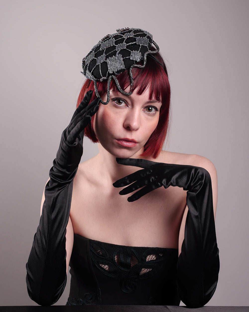 The model showcases the front of the Nova Pillbox Fascinator is an intricate constellation of geometry and texture. Its classic pillbox base in black wool felt is overlaid with a meticulously hand-stitched web of silver thread, creating a dimension by adding the formed rhinestone pipe grid that breaks into flowing strands at the edges. Designed by Merve Bayindir.