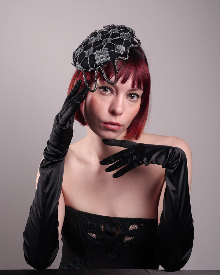 The model showcases the front of the Nova Pillbox Fascinator is an intricate constellation of geometry and texture. Its classic pillbox base in black wool felt is overlaid with a meticulously hand-stitched web of silver thread, creating a dimension by adding the formed rhinestone pipe grid that breaks into flowing strands at the edges. Designed by Merve Bayindir.