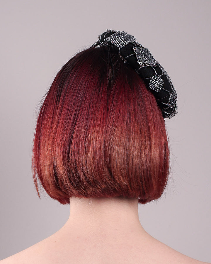 The model showcases the back of the Nova Pillbox Fascinator is an intricate constellation of geometry and texture. Its classic pillbox base in black wool felt is overlaid with a meticulously hand-stitched web of silver thread, creating a dimension by adding the formed rhinestone pipe grid that breaks into flowing strands at the edges. Designed by Merve Bayindir.