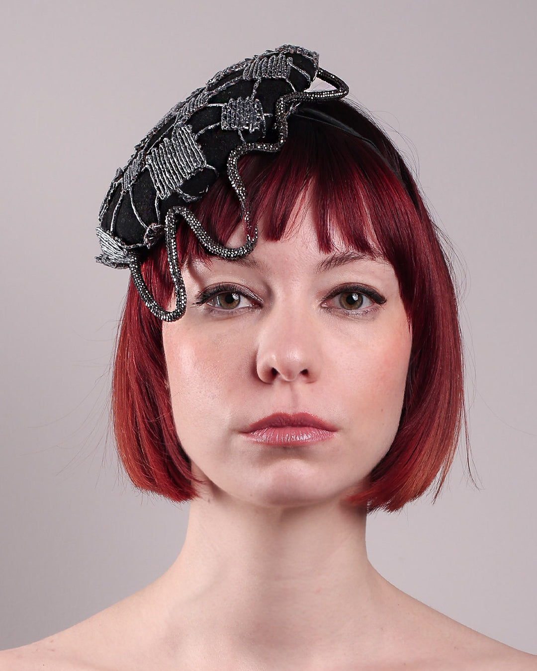The model showcases the front close up of the Nova Pillbox Fascinator is an intricate constellation of geometry and texture. Its classic pillbox base in black wool felt is overlaid with a meticulously hand-stitched web of silver thread, creating a dimension by adding the formed rhinestone pipe grid that breaks into flowing strands at the edges. Designed by Merve Bayindir.