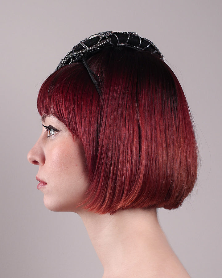 The model showcases the left side close up of the Nova Pillbox Fascinator is an intricate constellation of geometry and texture. Its classic pillbox base in black wool felt is overlaid with a meticulously hand-stitched web of silver thread, creating a dimension by adding the formed rhinestone pipe grid that breaks into flowing strands at the edges. Designed by Merve Bayindir.
