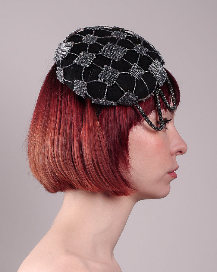 The model showcases the right close up of the Nova Pillbox Fascinator is an intricate constellation of geometry and texture. Its classic pillbox base in black wool felt is overlaid with a meticulously hand-stitched web of silver thread, creating a dimension by adding the formed rhinestone pipe grid that breaks into flowing strands at the edges. Designed by Merve Bayindir.