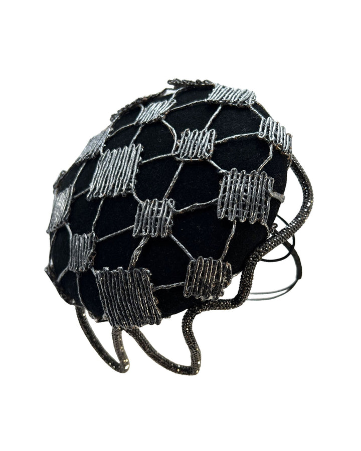 Still life picture of the Nova Pillbox Fascinator is an intricate constellation of geometry and texture. Its classic pillbox base in black wool felt is overlaid with a meticulously hand-stitched web of silver thread, creating a dimension by adding the formed rhinestone pipe grid that breaks into flowing strands at the edges. Designed by Merve Bayindir.