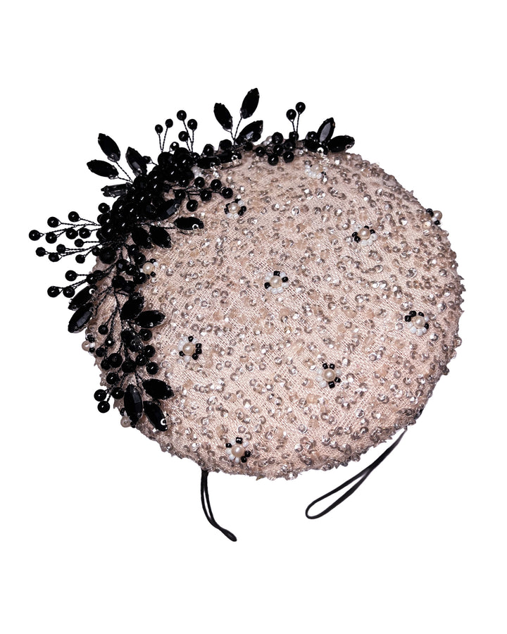 Blush sinamay fascinator with a rounded high-back silhouette, intricately embellished with clear crystals and pearl accents, and finished with a dramatic black beaded floral spray for elegant contrast. Designed and made by Merve Bayindir Millinery.

