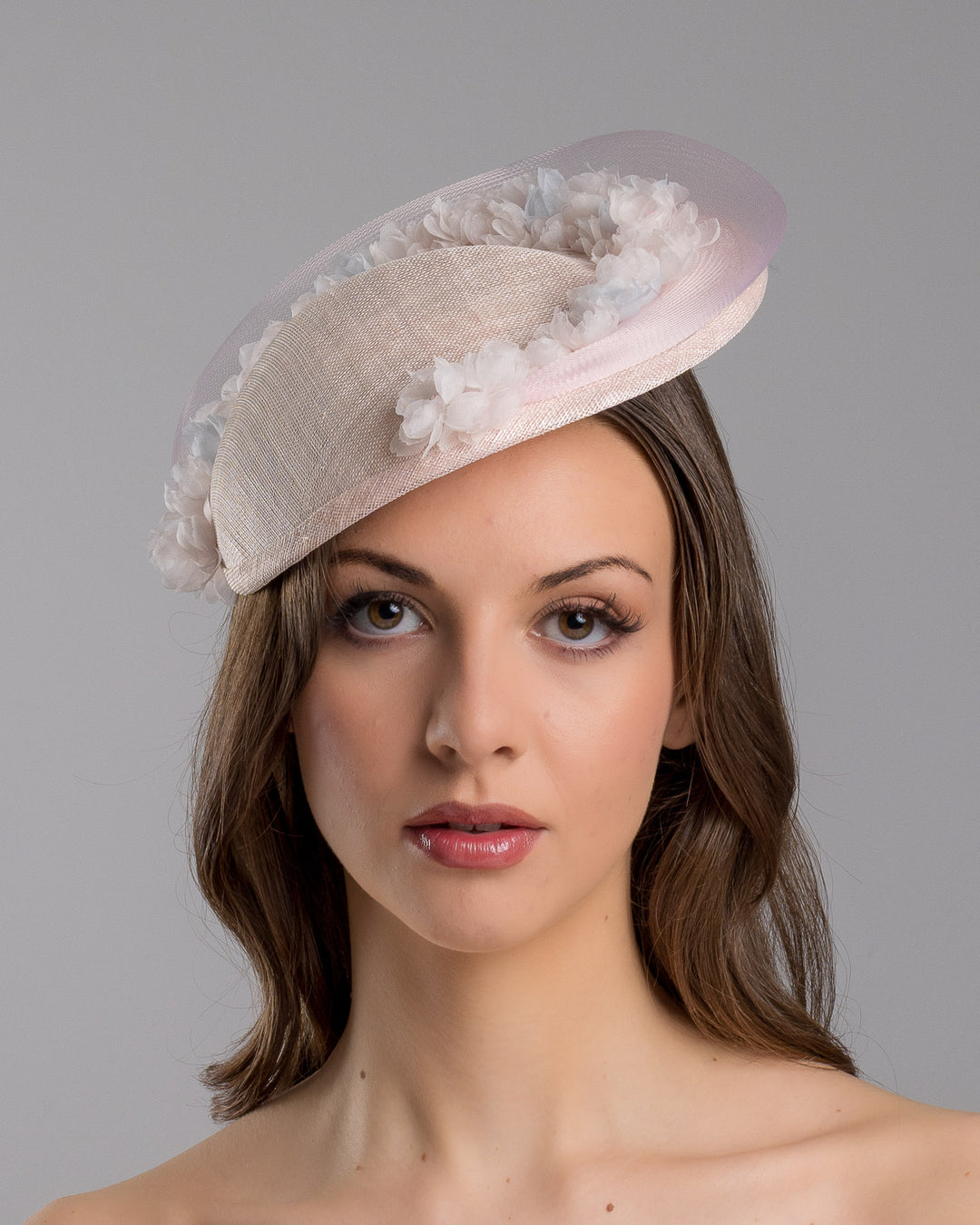 The model is showcasing the front side close up of the blush pink sinamay fascinator with a soft oval crown, trimmed with delicate pastel chiffon flowers and finished with a sheer pale veil overlay, mounted on a slim black headband. Designed and made by Merve Bayindir Millinery.
