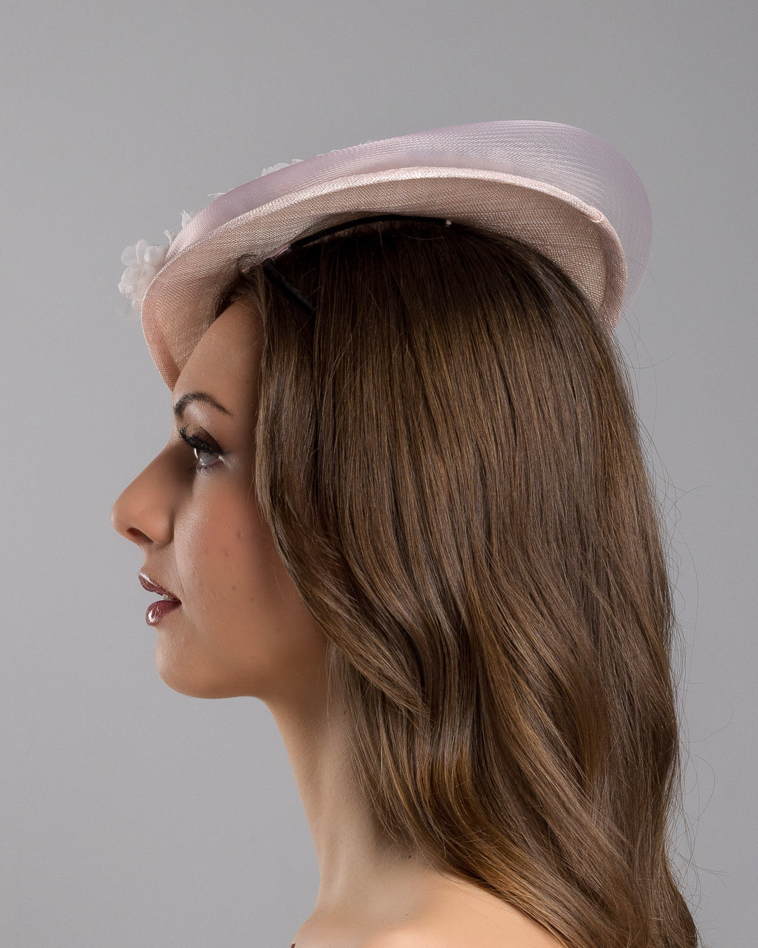 The model is showcasing the left side close up of the blush pink sinamay fascinator with a soft oval crown, trimmed with delicate pastel chiffon flowers and finished with a sheer pale veil overlay, mounted on a slim black headband. Designed and made by Merve Bayindir Millinery.
