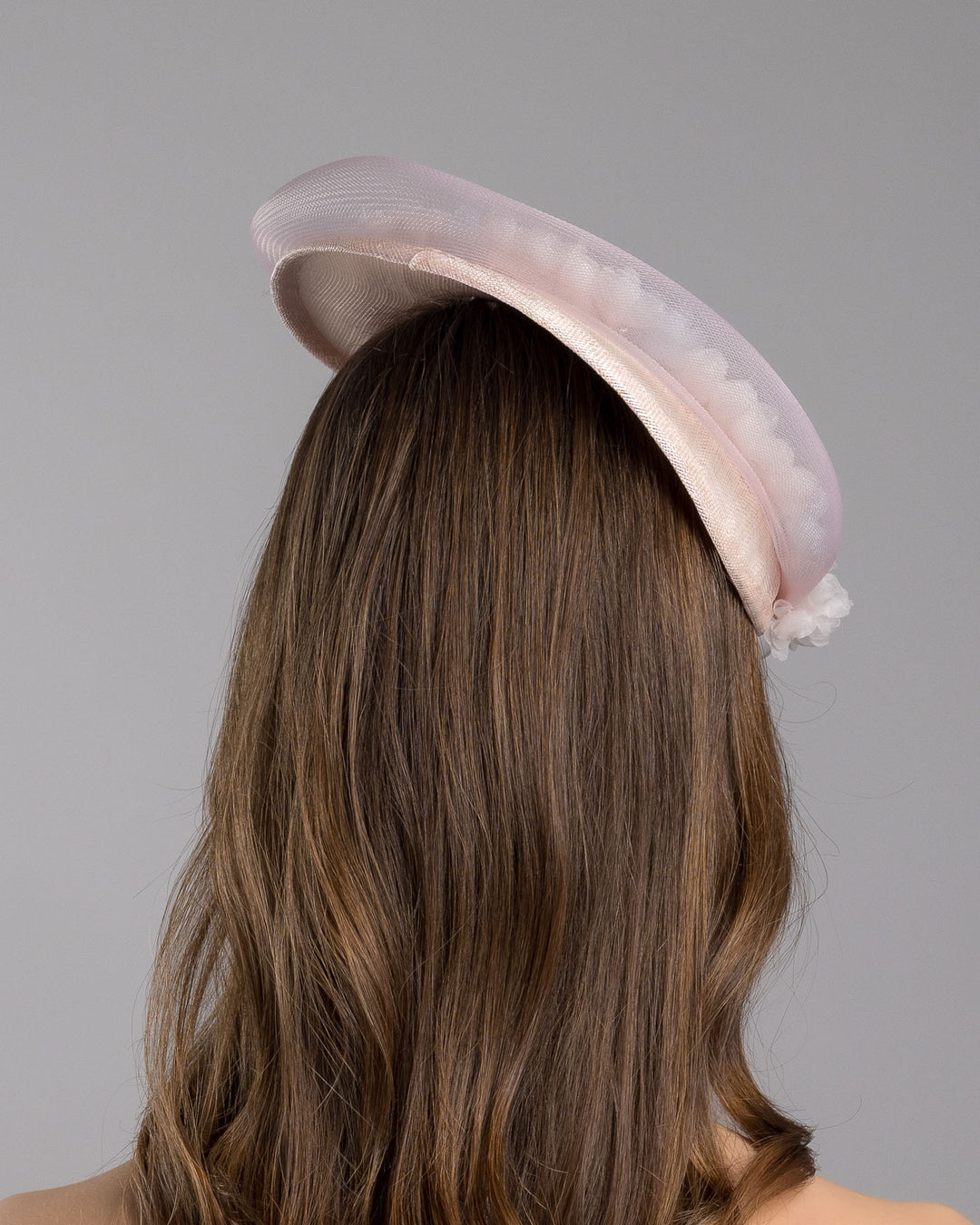The model is showcasing the back side close up of the blush pink sinamay fascinator with a soft oval crown, trimmed with delicate pastel chiffon flowers and finished with a sheer pale veil overlay, mounted on a slim black headband. Designed and made by Merve Bayindir Millinery.
