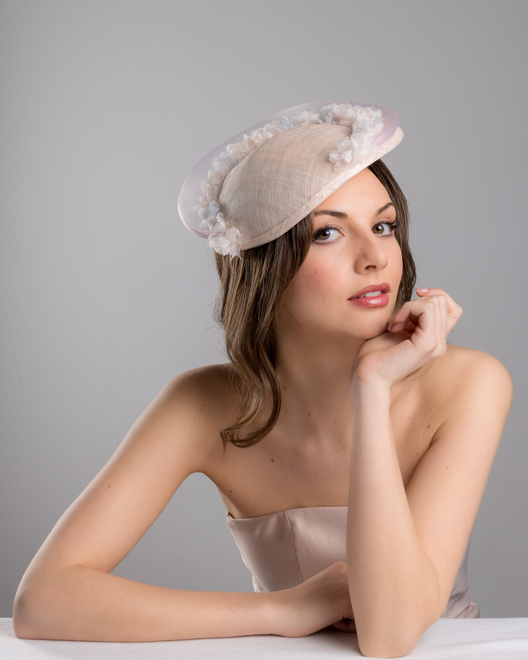 The female model is posing, wearing
Ollie, the blush pink sinamay fascinator with a soft oval crown, trimmed with delicate pastel chiffon flowers and finished with a sheer pale veil overlay, mounted on a slim black headband. Designed and made by Merve Bayindir Millinery.
