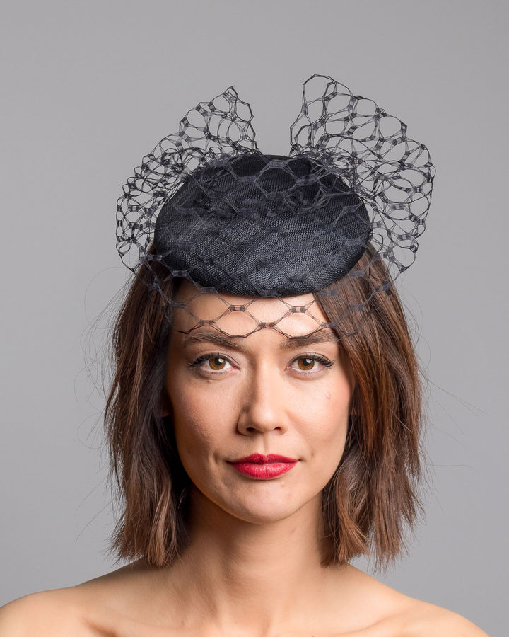 The model is showcasing the front side close up of the black round fascinator crafted from sinamay woven material, topped with sculpted looped detailing and framed by a dramatic open birdcage veil that flares outward on both sides. Designed and made by Merve Bayindir Millinery.
