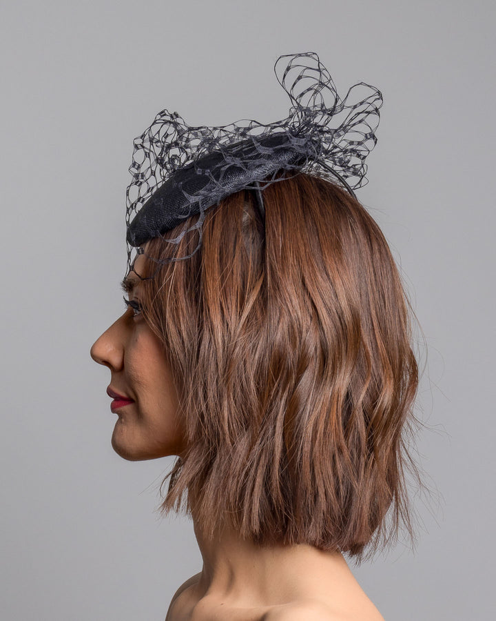 The model is showcasing the left side close up of the black round fascinator crafted from sinamay woven material, topped with sculpted looped detailing and framed by a dramatic open birdcage veil that flares outward on both sides. Designed and made by Merve Bayindir Millinery.
