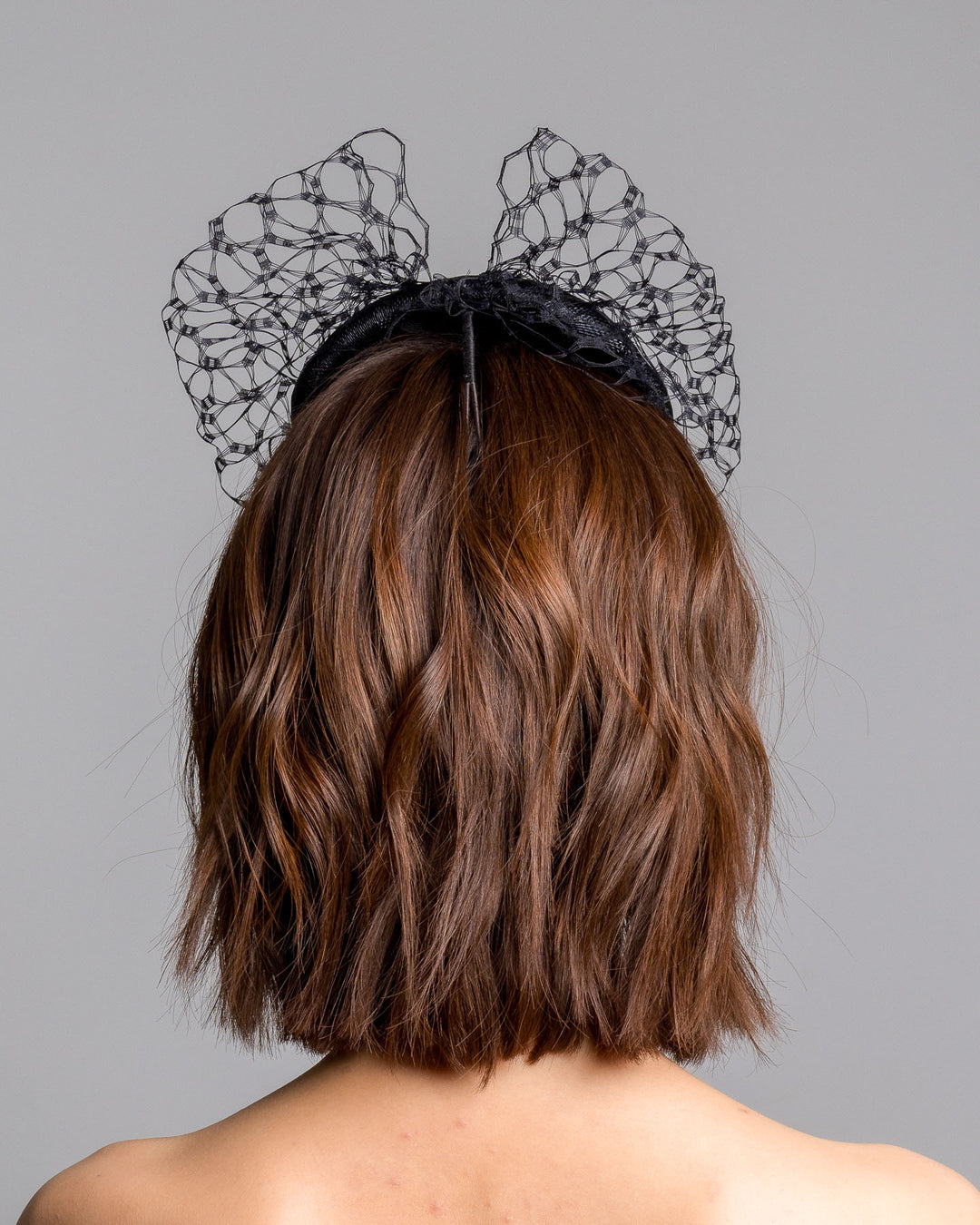 The model is showcasing the back side close up of the black round fascinator crafted from sinamay woven material, topped with sculpted looped detailing and framed by a dramatic open birdcage veil that flares outward on both sides. Designed and made by Merve Bayindir Millinery.
