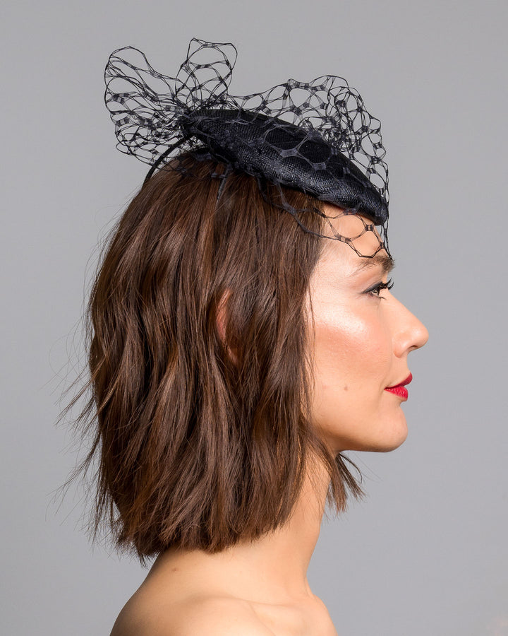 The model is showcasing the right side close up of the black round fascinator crafted from sinamay woven material, topped with sculpted looped detailing and framed by a dramatic open birdcage veil that flares outward on both sides. Designed and made by Merve Bayindir Millinery.
