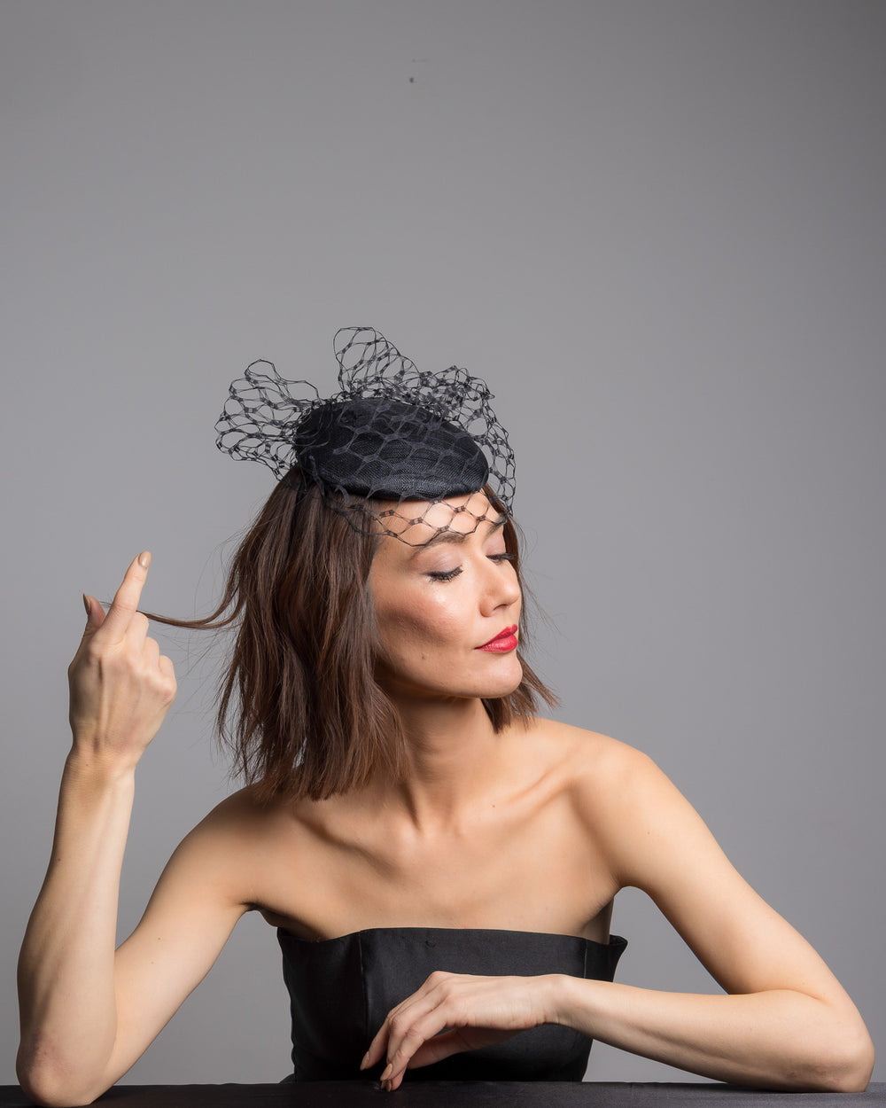 The female model is posing, wearing Ombra, the black round fascinator crafted from sinamay woven material, topped with sculpted looped detailing and framed by a dramatic open birdcage veil that flares outward on both sides. Designed and made by Merve Bayindir Millinery.
