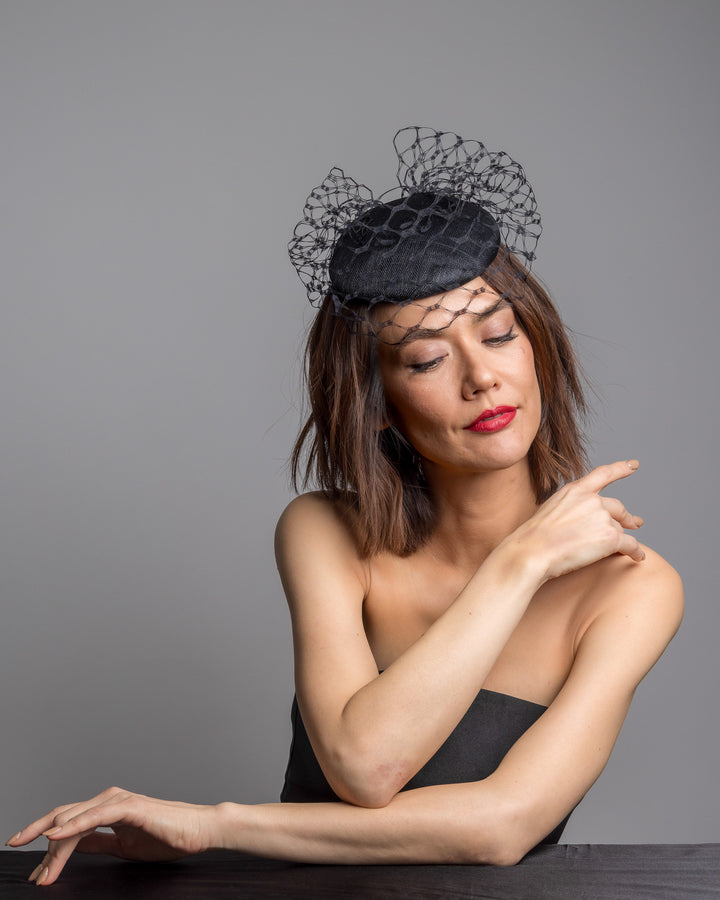 The female model is posing, wearing Ombra, the black round fascinator crafted from sinamay woven material, topped with sculpted looped detailing and framed by a dramatic open birdcage veil that flares outward on both sides. Designed and made by Merve Bayindir Millinery.
