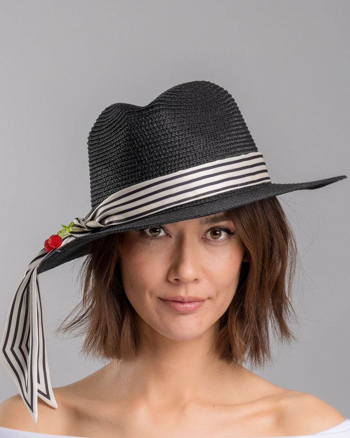 The model is showcasing the front side close up of the black woven straw boater hat with a wide brim, wrapped in a black and white striped ribbon and finished with long scarf ties accented by small red cherry embellishments. Designed and Made by Merve Bayindir millinery.