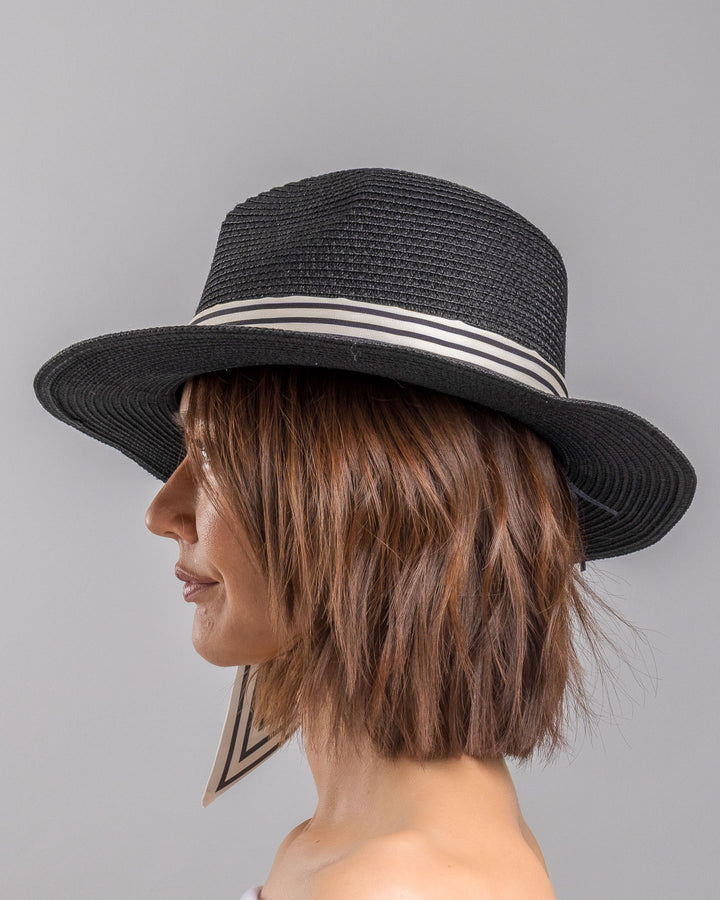 The model is showcasing the left side close up of the black woven straw boater hat with a wide brim, wrapped in a black and white striped ribbon and finished with long scarf ties accented by small red cherry embellishments. Designed and Made by Merve Bayindir millinery.