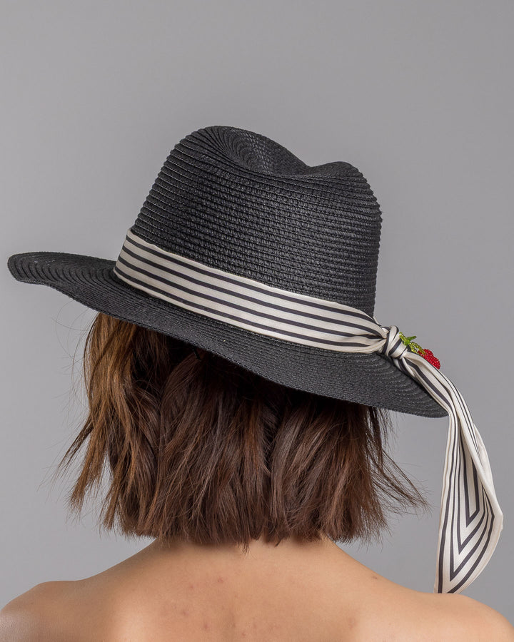 The model is showcasing the back side close up of the black woven straw boater hat with a wide brim, wrapped in a black and white striped ribbon and finished with long scarf ties accented by small red cherry embellishments. Designed and Made by Merve Bayindir millinery.