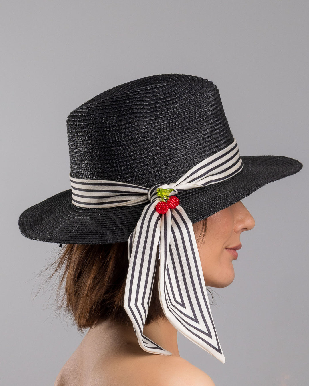 The model is showcasing the right side close up of the black woven straw boater hat with a wide brim, wrapped in a black and white striped ribbon and finished with long scarf ties accented by small red cherry embellishments. Designed and Made by Merve Bayindir millinery.