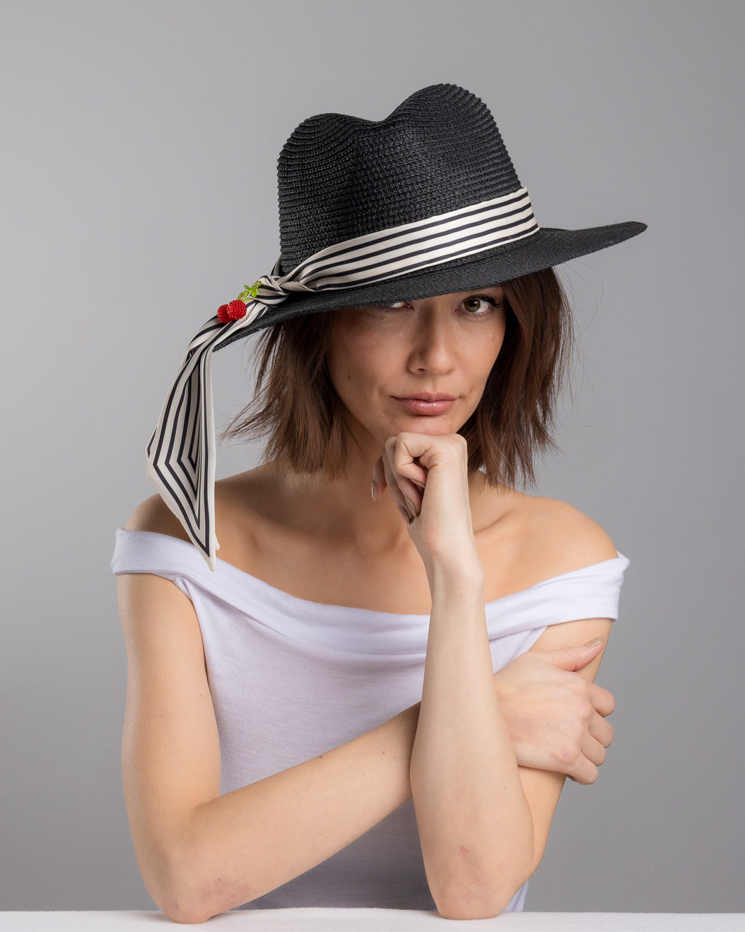 The female model is posing, wearing Onyxes, the black woven straw boater hat with a wide brim, wrapped in a black and white striped ribbon and finished with long scarf ties accented by small red cherry embellishments. Designed and Made by Merve Bayindir millinery.