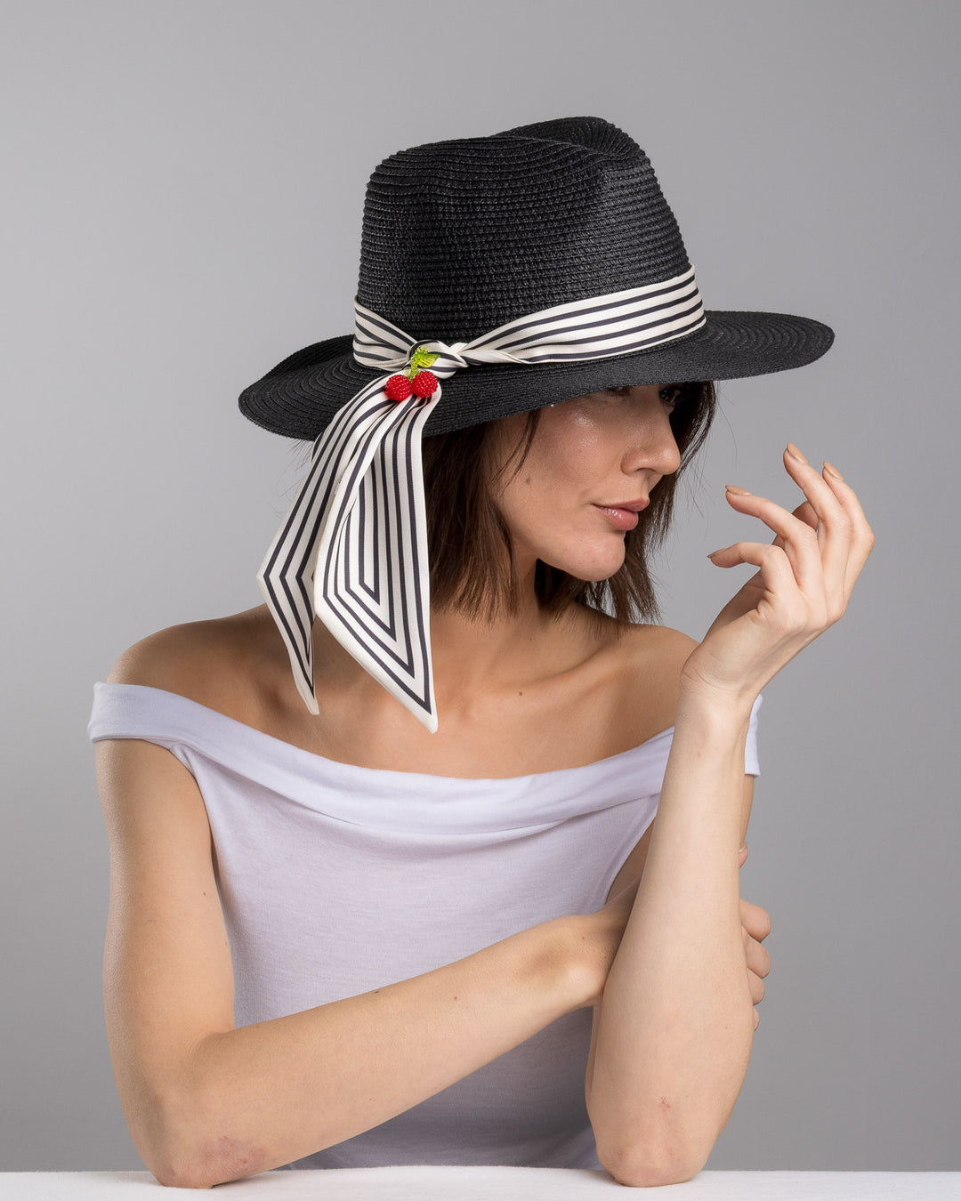 The female model is posing, wearing Onyxes, the black woven straw boater hat with a wide brim, wrapped in a black and white striped ribbon and finished with long scarf ties accented by small red cherry embellishments. Designed and Made by Merve Bayindir millinery.