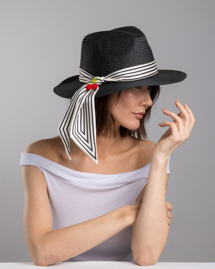 The female model is posing, wearing Onyxes, the black woven straw boater hat with a wide brim, wrapped in a black and white striped ribbon and finished with long scarf ties accented by small red cherry embellishments. Designed and Made by Merve Bayindir millinery.