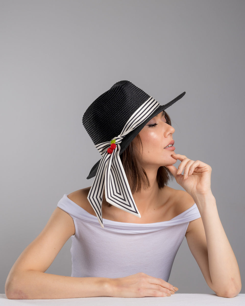 The female model is posing, wearing Onyxes, the black woven straw boater hat with a wide brim, wrapped in a black and white striped ribbon and finished with long scarf ties accented by small red cherry embellishments. Designed and Made by Merve Bayindir millinery.