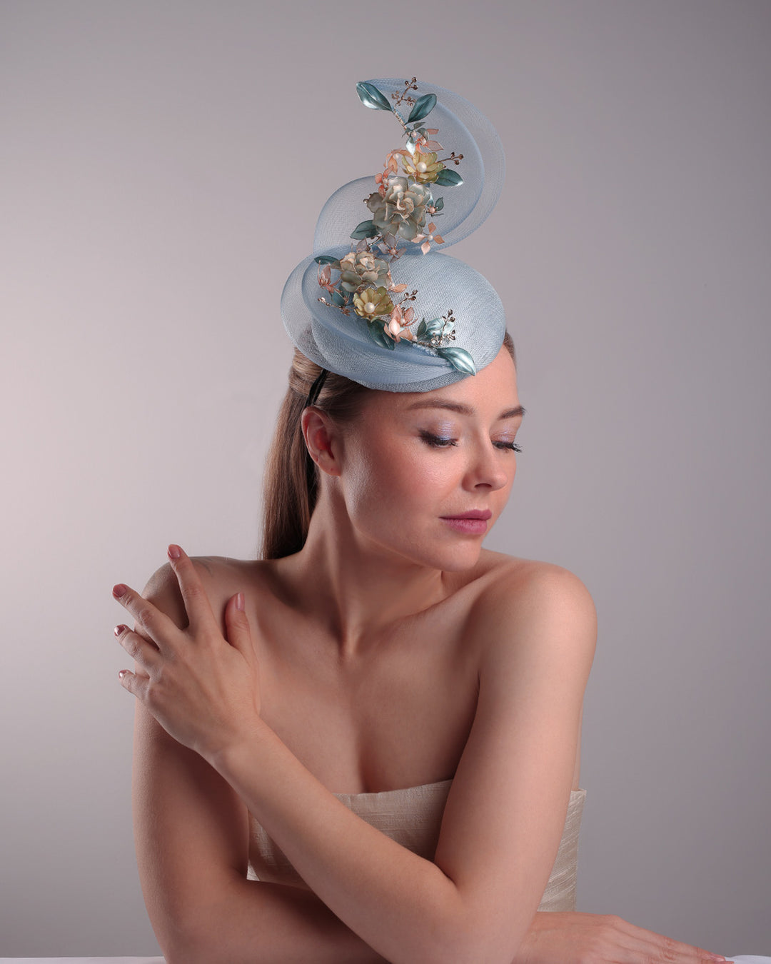 Model is showcasing the right front side of  a blue button shaped fasincator fitted with a wire headband and elastic. The button shaped fascinator is decorated with wave like crionline double swirl embellished with wired resin flower arrangments.
