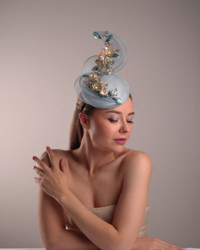 Model is showcasing the right front side of  a blue button shaped fasincator fitted with a wire headband and elastic. The button shaped fascinator is decorated with wave like crionline double swirl embellished with wired resin flower arrangments.