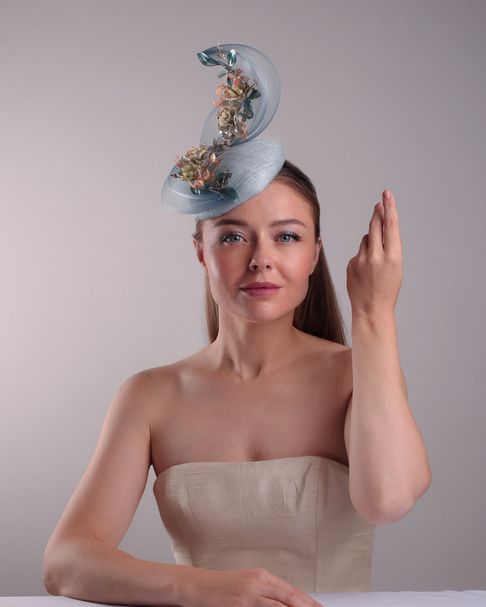 Model is showcasing the front of  a blue button shaped fasincator fitted with a wire headband and elastic. The button shaped fascinator is decorated with wave like crionline double swirl embellished with wired resin flower arrangments.