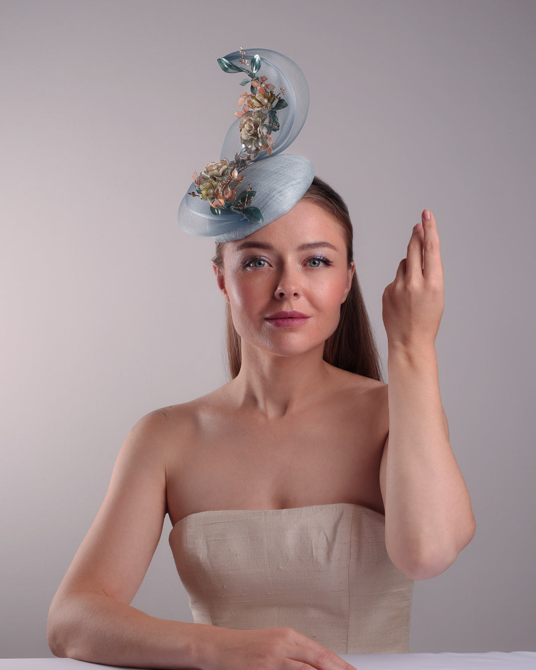 Model is showcasing the front of  a blue button shaped fasincator fitted with a wire headband and elastic. The button shaped fascinator is decorated with wave like crionline double swirl embellished with wired resin flower arrangments.