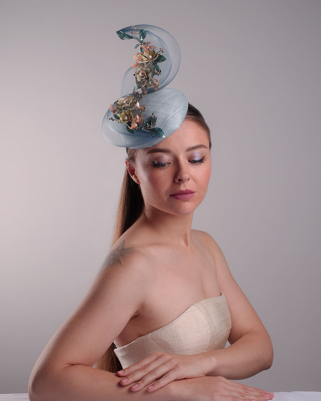 Model is showcasing the front of  a blue button shaped fasincator fitted with a wire headband and elastic. The button shaped fascinator is decorated with wave like crionline double swirl embellished with wired resin flower arrangments.