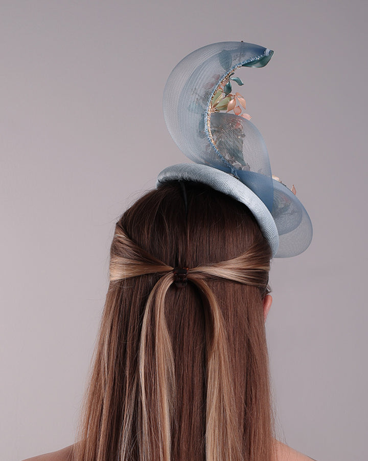 Model is showcasing the back side of  a blue button shaped fasincator fitted with a wire headband and elastic. The button shaped fascinator is decorated with wave like crionline double swirl embellished with wired resin flower arrangments.
