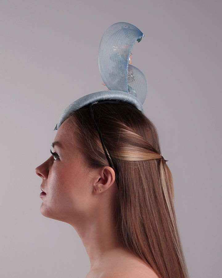 Model is showcasing the left side of  a blue button shaped fasincator fitted with a wire headband and elastic. The button shaped fascinator is decorated with wave like crionline double swirl embellished with wired resin flower arrangments.