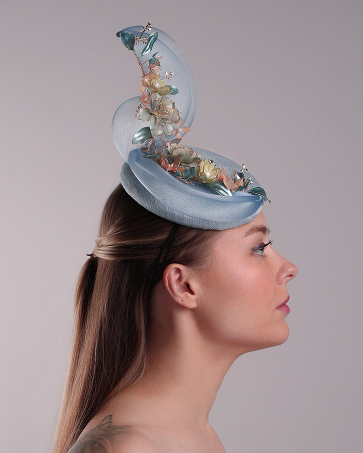 Model is showcasing the right side of  a blue button shaped fasincator fitted with a wire headband and elastic. The button shaped fascinator is decorated with wave like crionline double swirl embellished with wired resin flower arrangments.