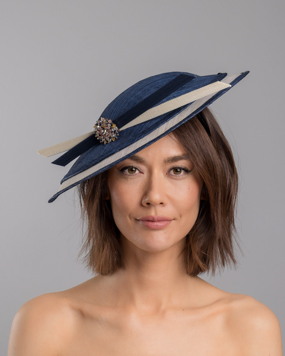 Model is showcasing the front side close up of multi layered tear drop shaped saucer in navy and white. This wide brim hat is decorated with navy and white buntal stripes and multi coloured crystal embellishments on the front. Designed and made by Merve Bayindir millinery.