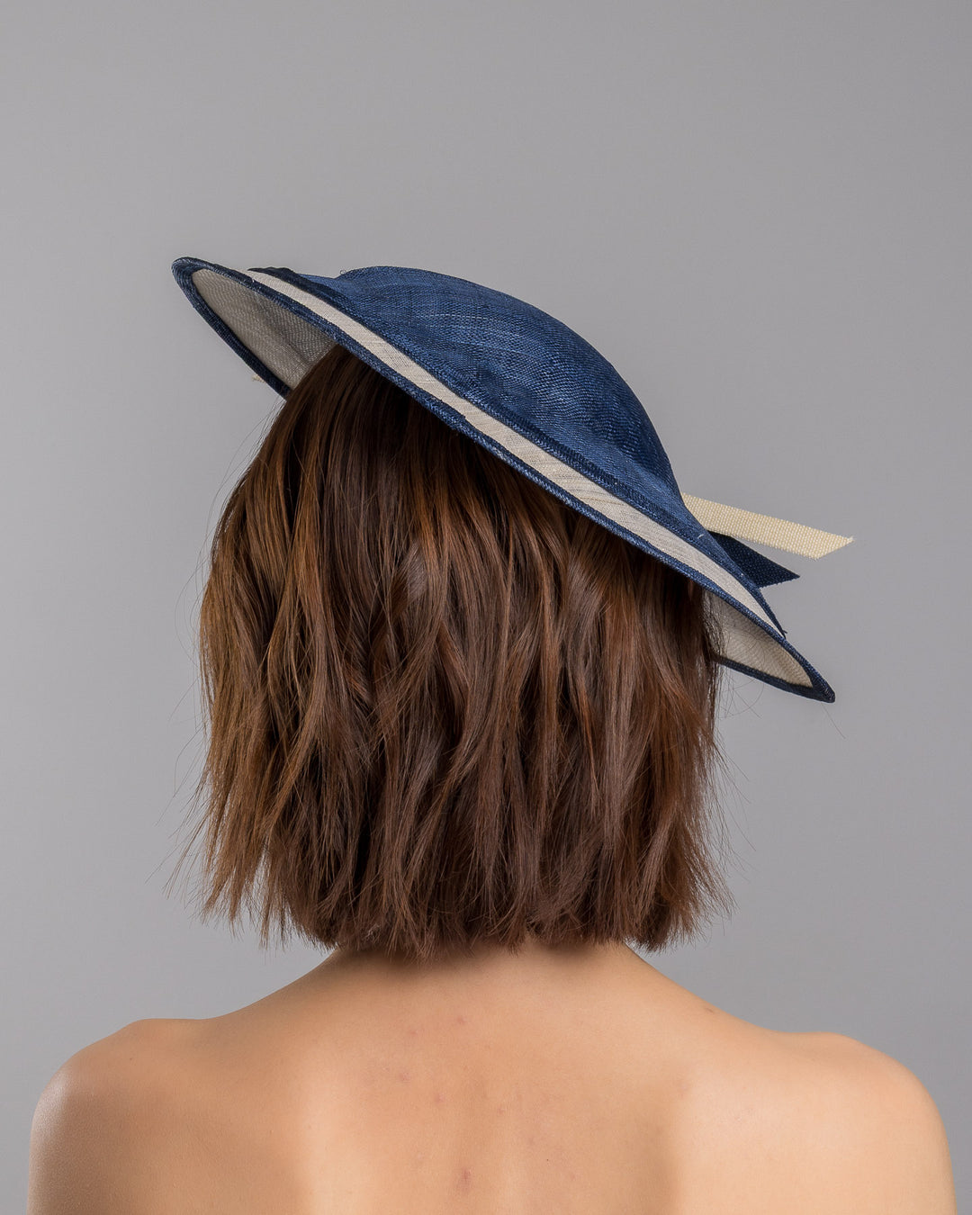 Model is showcasing the back side close up of multi layered tear drop shaped saucer in navy and white. This wide brim hat is decorated with navy and white buntal stripes and multi coloured crystal embellishments on the front. Designed and made by Merve Bayindir millinery.