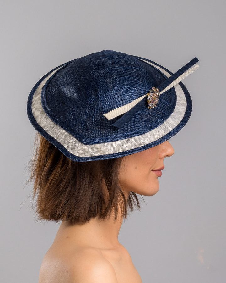 Model is showcasing the right side close up of multi layered tear drop shaped saucer in navy and white. This wide brim hat is decorated with navy and white buntal stripes and multi coloured crystal embellishments on the front. Designed and made by Merve Bayindir millinery.