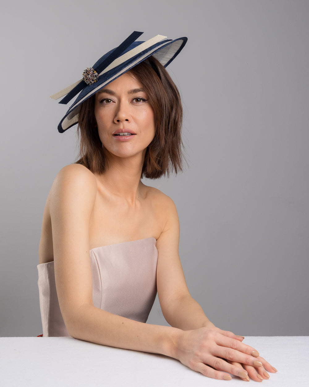 Model is posing wearing Ocean, multi layered tear drop shaped saucer in navy and white. This wide brim hat is decorated with navy and white buntal stripes and multi coloured crystal embellishments on the front. Designed and made by Merve Bayindir millinery.
