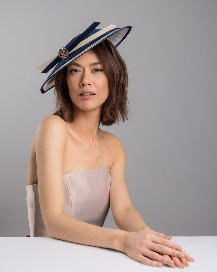 Model is posing wearing Ocean, multi layered tear drop shaped saucer in navy and white. This wide brim hat is decorated with navy and white buntal stripes and multi coloured crystal embellishments on the front. Designed and made by Merve Bayindir millinery.