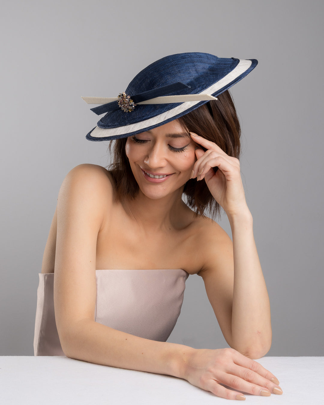 Model is posing wearing Ocean, multi layered tear drop shaped saucer in navy and white. This wide brim hat is decorated with navy and white buntal stripes and multi coloured crystal embellishments on the front. Designed and made by Merve Bayindir millinery.